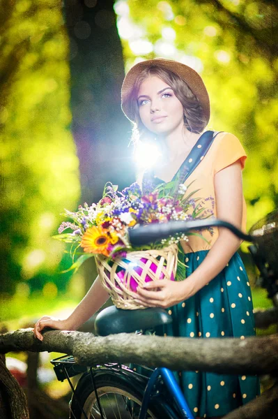 Bella ragazza con cappello carino e cesto con fiori divertirsi nel parco con la bicicletta. Sano concetto di stile di vita all'aperto. Paesaggio d'epoca. Bella ragazza bionda con look retrò con bici — Foto Stock