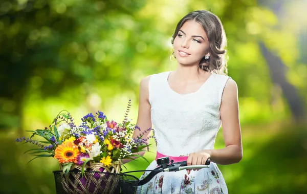 Beautiful girl wearing a nice white dress having fun in park with bicycle carrying a beautiful basket full of flowers. Vintage scenery. Pretty blonde girl with retro look, bike and basket with flowers Royalty Free Stock Images