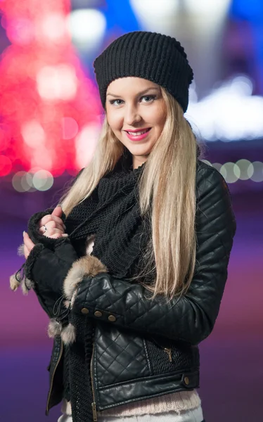 Fashionabla dam klädd i mössa och svart jacka utomhus i xmas landskap med blåljus i bakgrunden. porträtt av ung vacker kvinna med lång rättvis hår poserar leende i vinter stil. — Stockfoto