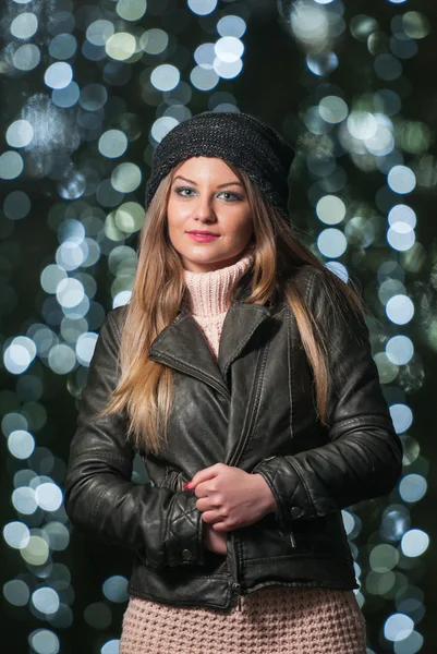 Fashionable lady wearing cap and black jacket outdoor in xmas scenery with blue lights in background. Portrait of young beautiful woman with long fair hair posing smiling in winter style. — Stock Photo, Image