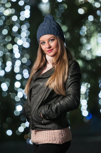 Fashionable lady wearing cap and black jacket outdoor in xmas scenery with blue lights in background. Portrait of young beautiful woman with long fair hair posing smiling in winter style. — Stock Photo, Image