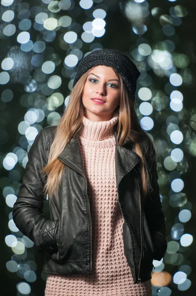 Donna alla moda che indossa berretto e giacca nera all'aperto in uno scenario natalizio con luci blu sullo sfondo. Ritratto di giovane bella donna con lunghi capelli biondi in posa sorridente in stile invernale . — Foto Stock