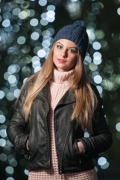 Fashionable lady wearing cap and black jacket outdoor in xmas scenery with blue lights in background. Portrait of young beautiful woman with long fair hair posing smiling in winter style. — Stock Photo, Image