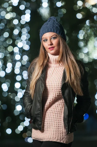 Fashionabla dam klädd i mössa och svart jacka utomhus i xmas landskap med blåljus i bakgrunden. porträtt av ung vacker kvinna med lång rättvis hår poserar leende i vinter stil. — Stockfoto