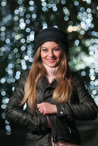 Modne damy sobie czapkę i czarna kurtka odkryty w Boże Narodzenie dekoracje z niebieskiego światła w tle. portret młodej kobiety piękne z długie jasne włosy, pozowanie, uśmiechając się w stylu zimy. — Zdjęcie stockowe