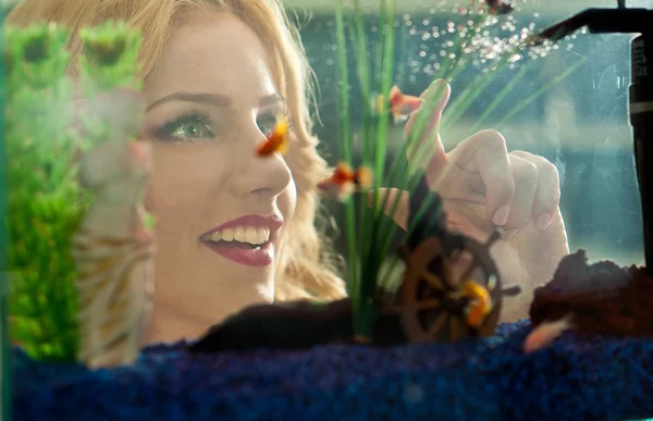 Hermosa chica rubia mirando peces dorados en el acuario. Atractiva hembra con una hermosa sonrisa admirando un gran acuario. Mujer bonita jugando con peces de colores nadando en el acuario —  Fotos de Stock