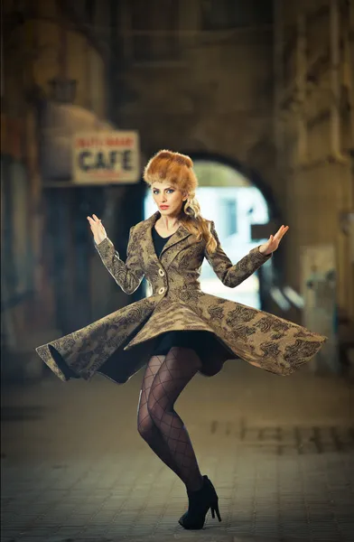 Attractive elegant blonde young woman wearing an outfit with Russian influence in urban fashion shot. Beautiful fashionable young girl with long legs and fur cap posing spinning on street — Stock Photo, Image