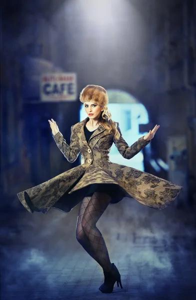 Attractive elegant blonde young woman wearing an outfit with Russian influence in urban fashion shot. Beautiful fashionable young girl with long legs and fur cap posing spinning on street — Stock Photo, Image