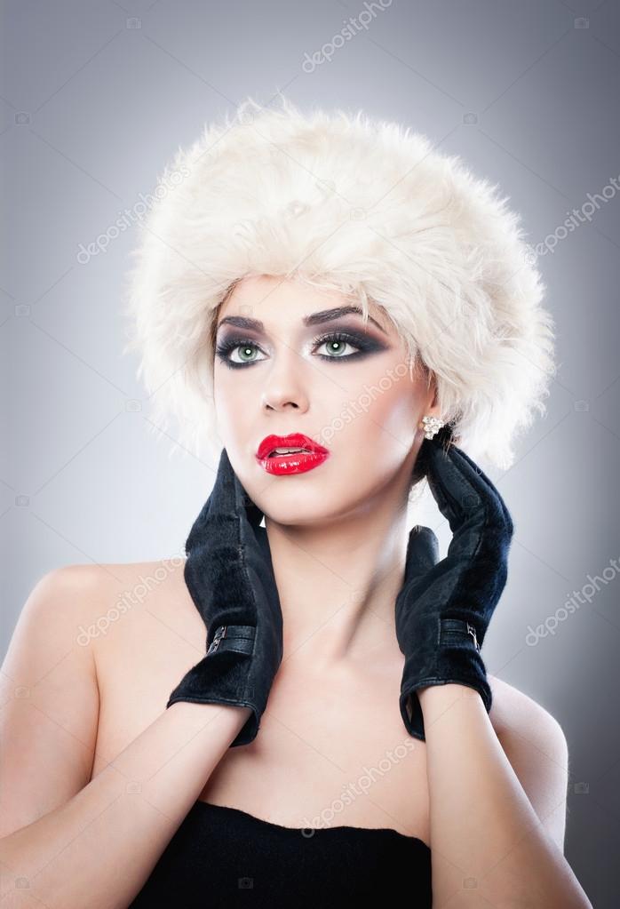 Attractive young Caucasian adult with black gloves isolated on grey background. Beautiful girl with red lips in white fur hat - studio isolated. Make up - beautiful female art portrait studio isolated