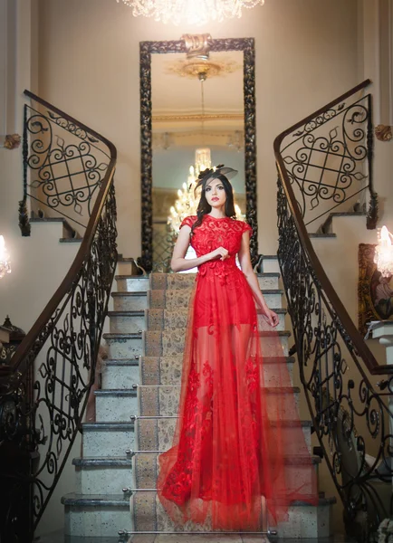 The beautiful girl in a long red dress posing in a vintage scene. Young beautiful woman wearing a red dress in an old hotel. Sensual elegant young woman in red long dress indoor shot. — Stock Photo, Image