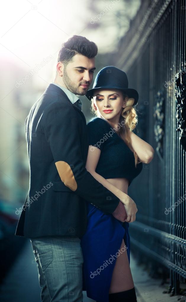 Couple in love in railway station. Beautiful well-dressed couple standing on railway platform. Handsome brunette young man holding a fashionable blonde with hat next to a train