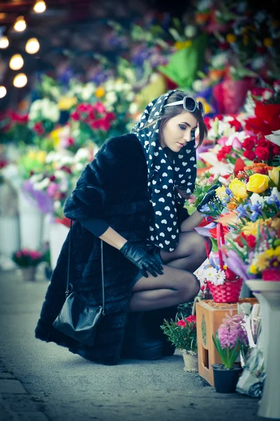 Beautiful brunette woman with gloves choosing flowers at the florist shop. Fashionable female with sunglasses and head scarf at flower shop. Pretty brunette in black choosing flowers - urban shot — Stock Photo, Image