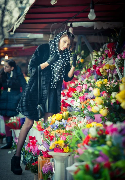 Beautiful brunette woman with gloves choosing flowers at the florist shop. Fashionable female with sunglasses and head scarf at flower shop. Pretty brunette in black choosing flowers - urban shot — Stock Photo, Image