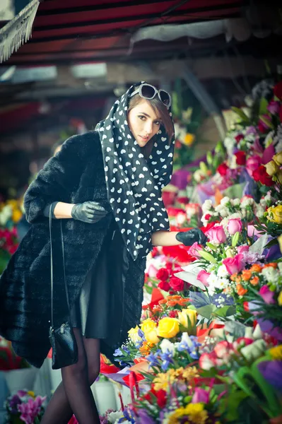 Hermosa mujer morena con guantes eligiendo flores en la floristería. Mujer de moda con gafas de sol y pañuelo en la tienda de flores. Bonita morena en negro eligiendo flores - tiro urbano —  Fotos de Stock