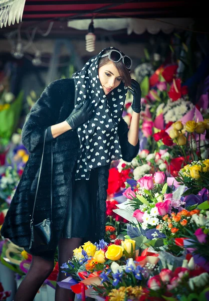 Beautiful brunette woman with gloves choosing flowers at the florist shop. Fashionable female with sunglasses and head scarf at flower shop. Pretty brunette in black choosing flowers - urban shot — Stock Photo, Image