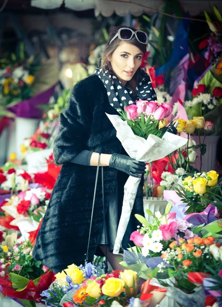 Belle femme brune avec des gants choisissant des fleurs à la boutique de fleuristes. Femme à la mode avec des lunettes de soleil et foulard au magasin de fleurs. Jolie brune au choix noir fleurs - plan urbain — Photo