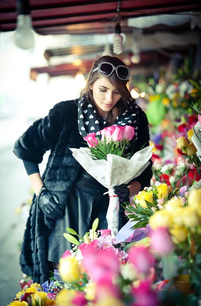 Belle femme brune avec des gants choisissant des fleurs à la boutique de fleuristes. Femme à la mode avec des lunettes de soleil et foulard au magasin de fleurs. Jolie brune au choix noir fleurs - plan urbain — Photo