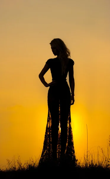 Silhouette di una giovane donna in abito lungo trasparente davanti a un bel tramonto. Bionda in pizzo nero in posa all'aperto davanti a un bellissimo tramonto. Ragazza sensuale con il sole che sorge dietro di lei . — Foto Stock