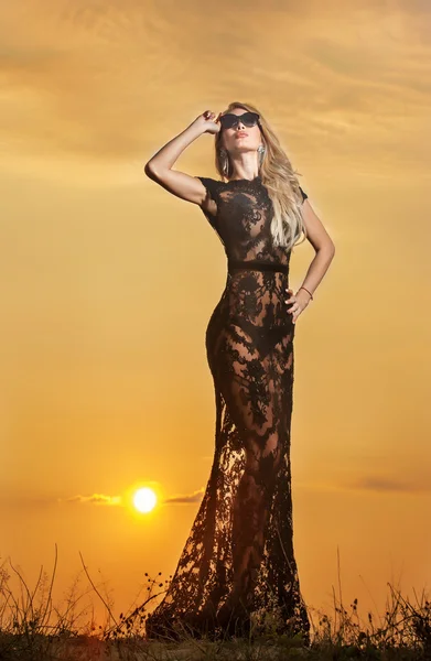 Silhouette einer jungen Frau in langem transparentem Kleid vor einem wunderschönen Sonnenuntergang. Blondine in schwarzem Spitzenkleid posiert draußen vor einem wunderschönen Sonnenuntergang. sinnliches Mädchen mit aufgehender Sonne hinter sich. — Stockfoto