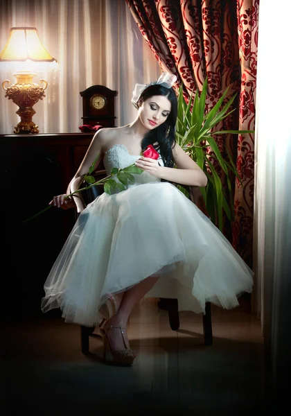 Young beautiful luxurious bride sitting holding a red rose in classic scenery. Attractive bride with curtains and lamp in background. Seductive brunette with ribbon in luxury manor, vintage style — Stock Photo, Image