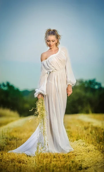 Young woman with long white dress standing on a wheat field. Portrait of girl outdoor. Romantic young woman posing on clean blue sky. Attractive woman in white dress in yellow wheat field at sunrise. — Stock Photo, Image