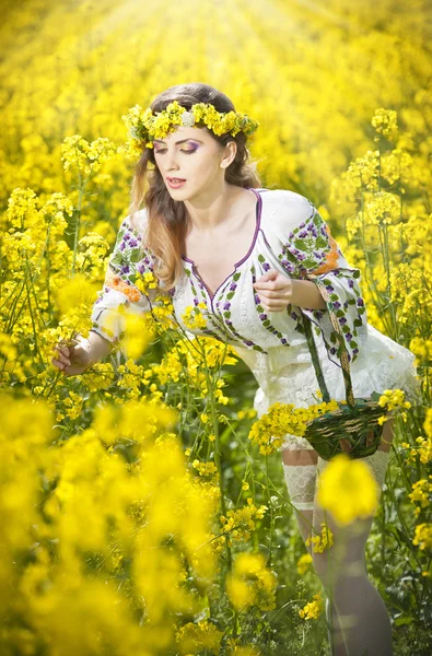 Young girl wearing Romanian traditional blouse holding a basket in canola field, outdoor shot. Portrait of beautiful blonde with blue eyes smiling and enjoying the bright yellow flowers of rapeseed — Stock Photo, Image