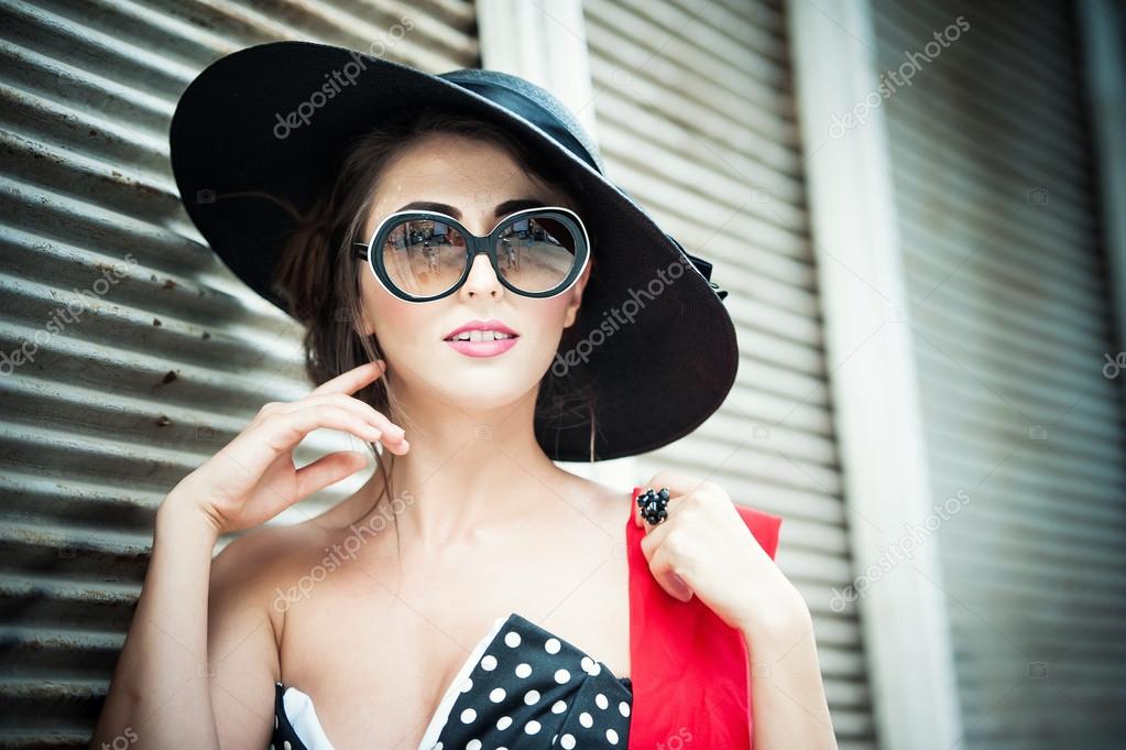 Attractive brunette girl with black hat, red scarf and sunglasses posing outdoor. Beautiful fashionable young woman with modern accessories, urban shot. Gorgeous brunette with large black hat smiling.
