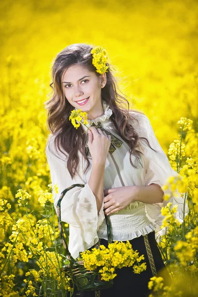 Pretty long hair girl wearing white blouse posing holding a basket in canola field, outdoor shot. Portrait of beautiful brunette with bright yellow flowers in hair smiling and enjoying the rapeseed — Stock Photo, Image