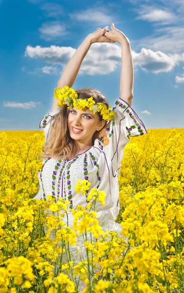 Giovane ragazza che indossa camicetta tradizionale rumena in posa in campo di colza con cielo nuvoloso in background, tiro all'aperto. Ritratto di bella bionda con ghirlanda di fiori sorridente nel campo di colza — Foto Stock