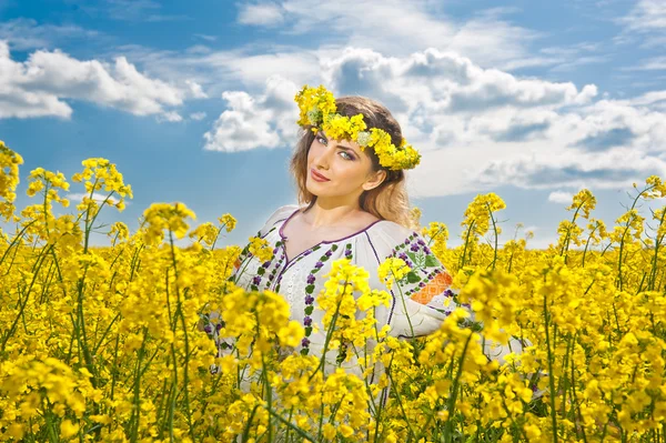 Chica joven con la blusa tradicional rumana posando en el campo de canola con el cielo nublado en el fondo, tiro al aire libre. Retrato de hermosa rubia con flores corona sonriendo en campo de colza —  Fotos de Stock