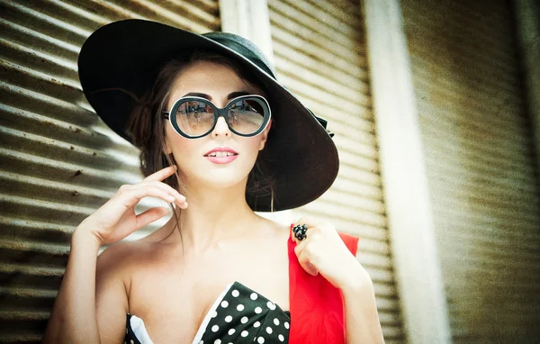 Attraente ragazza bruna con cappello nero, sciarpa rossa e occhiali da sole in posa all'aperto. Bella giovane donna alla moda con accessori moderni, colpo urbano. Splendida bruna con grande cappello nero sorridente . — Foto Stock