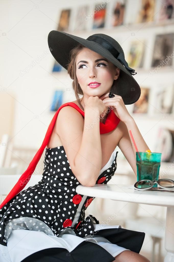 Fashionable attractive lady with black hat and red scarf sitting on chair in restaurant, indoor shot. Young woman posing in elegant scenery. Art photo of elegant sensual woman, vintage style