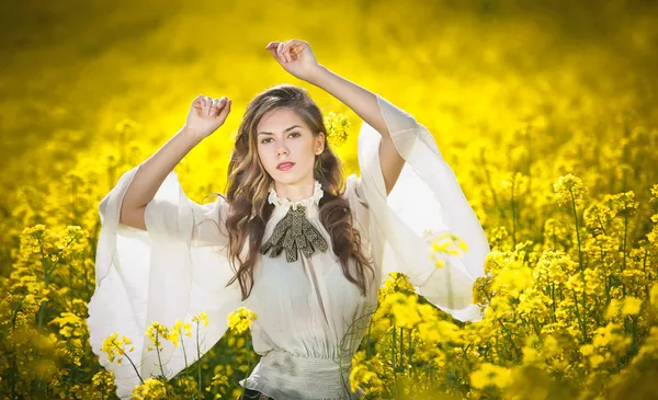 Junges Mädchen in eleganter weißer Bluse posiert im Rapsfeld, Außenaufnahme. Porträt der schönen langen Haare brünett mit großen transparenten Ärmeln in leuchtend gelben Rapsfeld, Frühlingslandschaft — Stockfoto