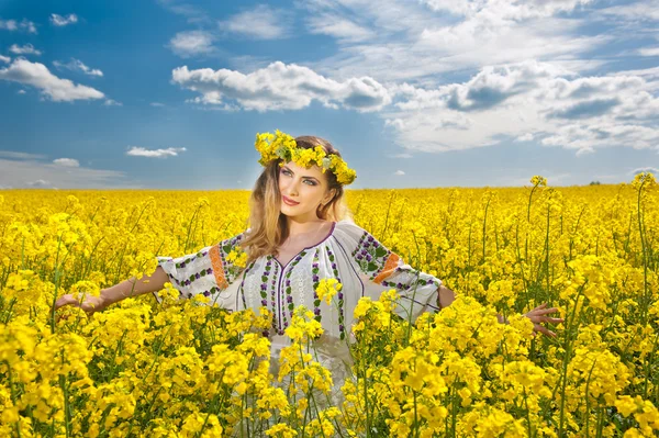 Mladá dívka, která nosí rumunské tradiční halenka pózuje v poli řepky s zatažené obloze v pozadí, venkovní zastřelen. portrét krásná blondýnka s květy věnec s úsměvem v řepkovém poli — Stock fotografie