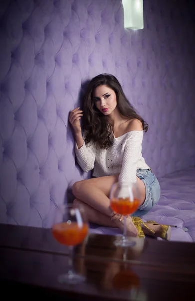 Beautiful brunette girl in denim shorts and white blouse posing  on lilac textured background with two drink glasses foreground. Young sensual woman siting alone in a modern night club. — Stock Photo, Image