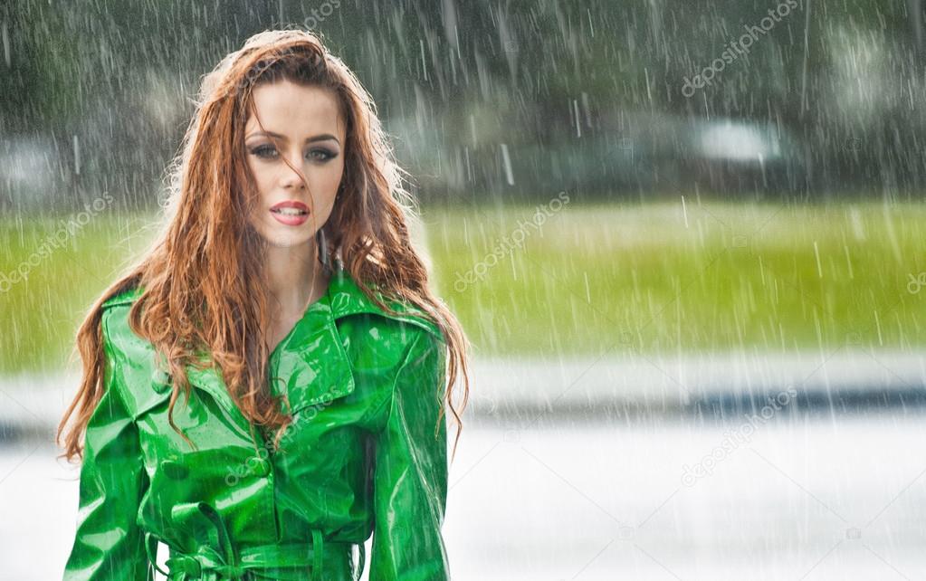 Beautiful woman in bright green coat posing in the rain. Dramatic redhead staying in the rain drops, urban shot. Attractive red hair girl on the street in a rainy day. Emotional pretty young female.