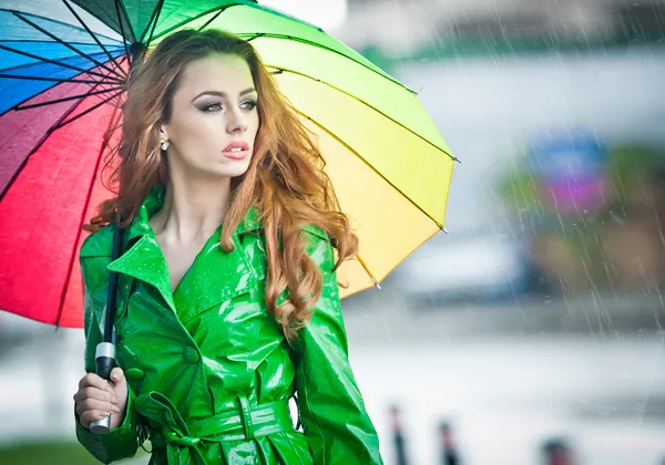 Beautiful woman in bright green coat posing in the rain holding a multicolored umbrella. Dramatic redhead staying under umbrella, urban shot. Attractive red hair girl on the street in a rainy day. — Stock Photo, Image