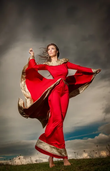 Modische schöne junge Frau im roten langen Kleid posiert im Freien mit bewölkten dramatischen Himmel im Hintergrund. attraktive lange Haare brünettes Mädchen mit elegantem luxuriösen Kleid am stürmischen Himmel. — Stockfoto