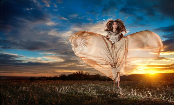 Modieuze mooie jonge vrouw in naakt gekleurde lange jurk spinnen rond op zoek als een vlinder op bewolkte dramatische hemel in de zonsondergang. aantrekkelijke brunette met elegante luxe jurk, buiten schot. — Stockfoto