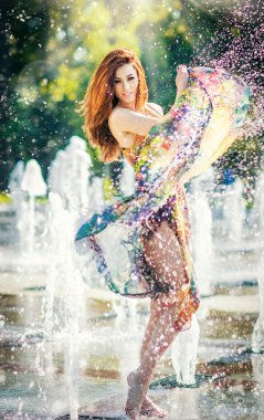 Attractive girl in multicolored short dress playing with water in a summer hottest day. Girl with wet dress enjoying fountains. Young beautiful happy female playing with outdoor water fountains.