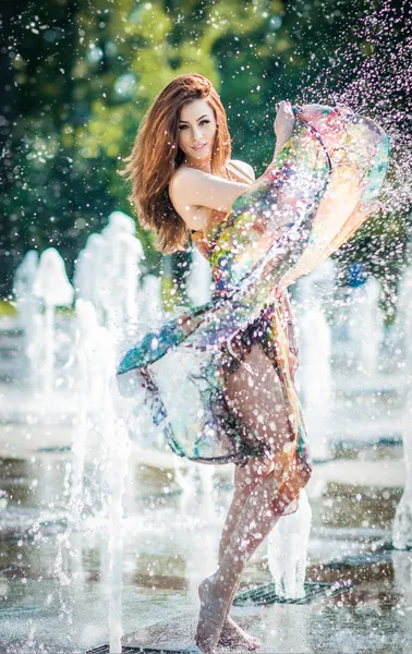 Jolie fille en robe courte multicolore jouant avec de l'eau dans une journée la plus chaude d'été. Fille avec robe humide profiter des fontaines. Jeune belle femme heureuse jouer avec des fontaines d'eau en plein air . — Photo