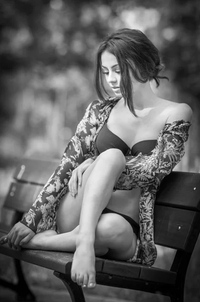 Attractive girl in swimsuit sitting relaxed on a bench. Fashionable female model with romantic look posing in park. Beautiful woman in bikini with nice legs sitting daydreaming, black and white. — Stock Photo, Image
