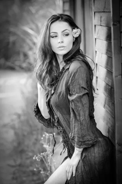 Fashion portrait of sexy brunette in black blouse leaning on wooden cabin wall. Sensual attractive woman with a flower in hair, black and white photo. Perfect body girl with long hair posing outdoor — Stock Photo, Image