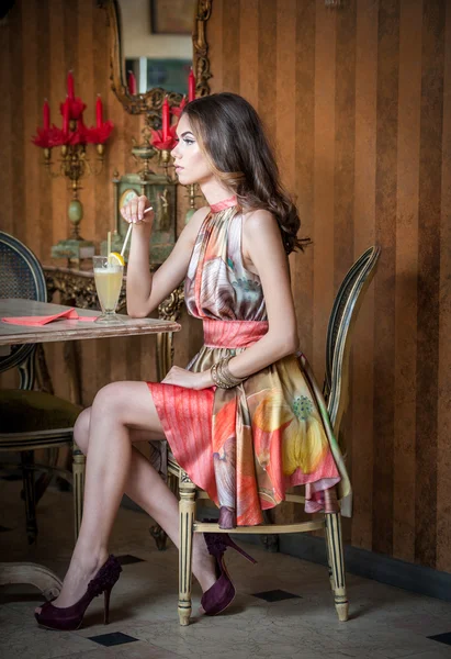 Mujer atractiva de moda en vestido multicolor sentado en el restaurante. Hermosa morena posando en un elegante paisaje vintage con un vaso de jugo. Atractiva hembra bebiendo limonada en la cafetería — Foto de Stock