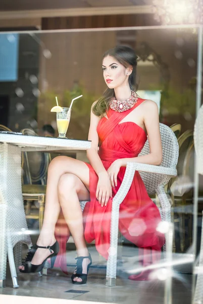 Modieuze aantrekkelijke jonge vrouw in rode jurk zittend in restaurant, buiten de ramen. mooie brunette poseren in elegante vintage landschap met een glas limonade. concept van de foto door het raam — Stockfoto