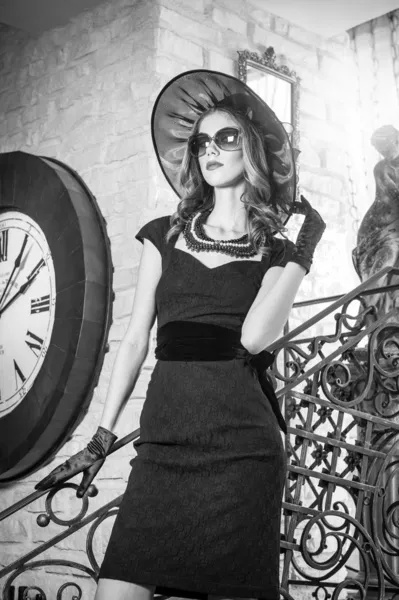 Young beautiful brunette woman in black standing on stairs near an over sized wall clock. Elegant romantic mysterious lady with movie star look in luxurious vintage interior, black and white photo. — Stock Photo, Image