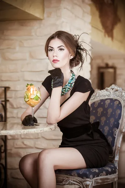 Mujer joven atractiva de moda en vestido negro sentado en el restaurante. Hermosa morena posando en un elegante paisaje vintage con un vaso de jugo. Señora atractiva con guantes en el interior de lujo —  Fotos de Stock