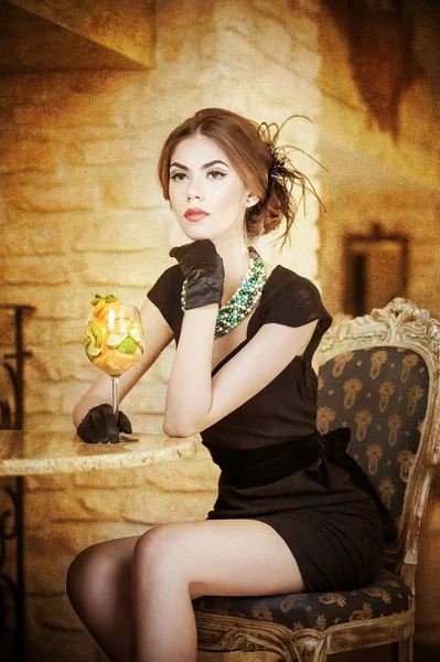 Mujer joven atractiva de moda en vestido negro sentado en el restaurante. Hermosa morena posando en un elegante paisaje vintage con un vaso de jugo. Señora atractiva con guantes en el interior de lujo — Foto de Stock