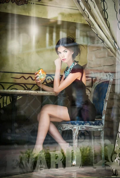 Modieuze aantrekkelijke jonge vrouw in zwarte jurk zittend in restaurant, buiten het venster. mooie brunette poseren in elegante vintage landschap met een glas sap. concept van de foto door het raam — Stockfoto