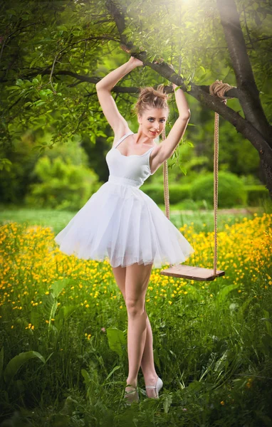 Muchacha atractiva en vestido corto blanco posando cerca de un columpio de árbol con un prado florido en el fondo. Mujer joven rubia con vestido de bailarina bajo un árbol en una posición de ballet durante un día de verano brillante . —  Fotos de Stock